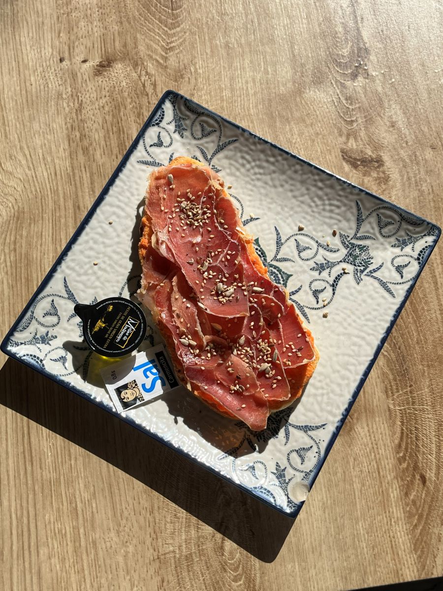 Tostada de jamón con tomate y sésamo sobre cerámica azul pintada a mano, bañada por la luz de la mañana.